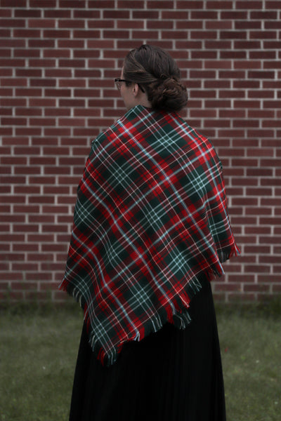 woman wearing a red and green plaid shawl facing a brick wall.