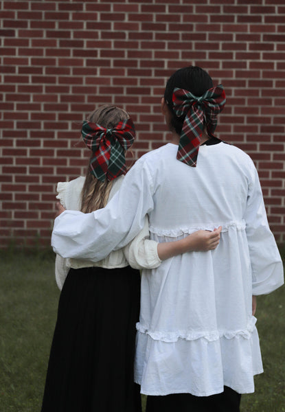 Two people wearing white blouses with plaid bows in their hair against a brick wall.