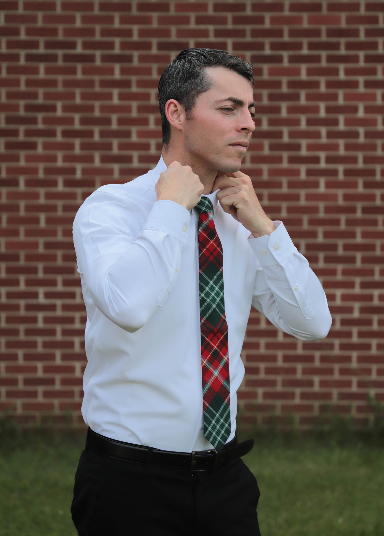 Man adjusting his red and green plaid tie against a brick wall.