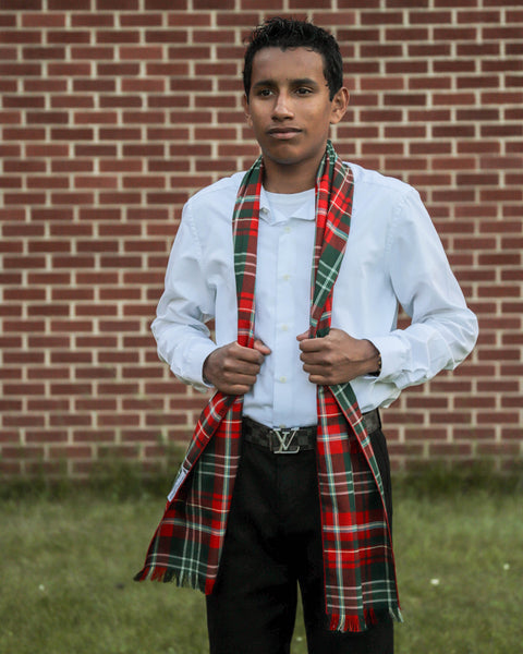 Youth wearing a red and green plaid scarf over a white shirit in front of a brick wall.