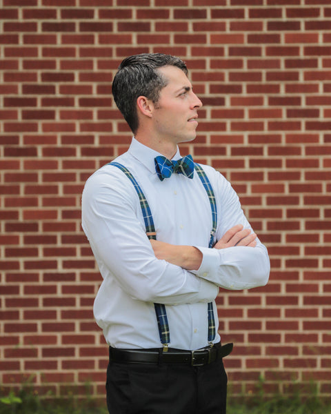 Man wearing a white shirt, blue bow tie, and plaid suspenders standing against a brick wall.