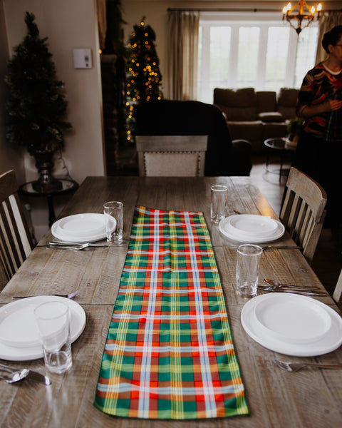 A festive holiday table runner with a green, red, yellow, and white plaid pattern Northwest Territories tartan laid down the center of a wooden dining table set with white plates and glasses in a cozy living room.