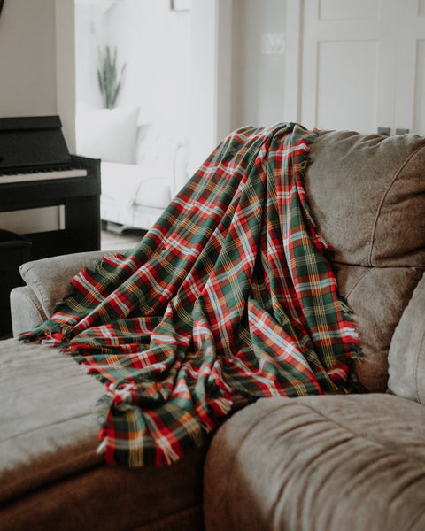 A red, green, and yellow plaid Northwest Territories tartan throw blanket is draped over a brown fabric armchair in a softly lit living room.
