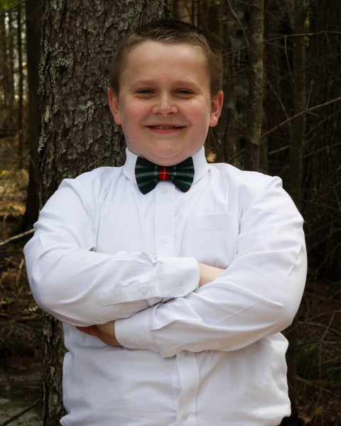 Young boy in a white shirt and a red green and yellow plaid bow tie standing in front of a tree.