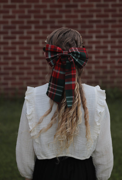 Person with plaid hair bow and white ruffled shirt against a brick wall.