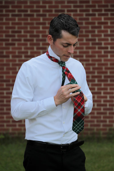 Man tying a New Brunswick plaid tie against a brick wall wearing a white shirt and black dress pants.