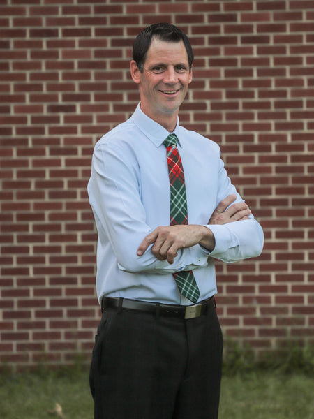 Man wearing a light blue shirt, dark pants, and a New Brunswick tartan plaid tie standing in front of a brick wall.