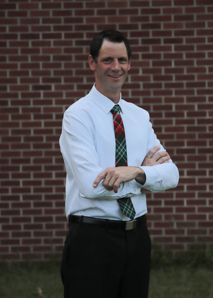 Man wearing a white shirt and green, red, and white plaid tie standing against a brick wall.
