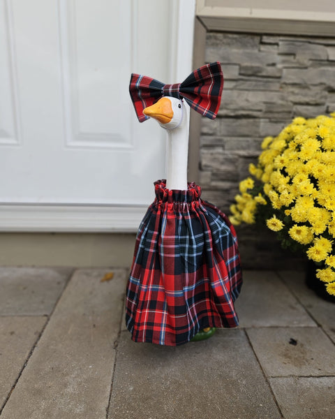 White cement goose with red and green plaid dress in front of white door, brick wall and yellow flowers