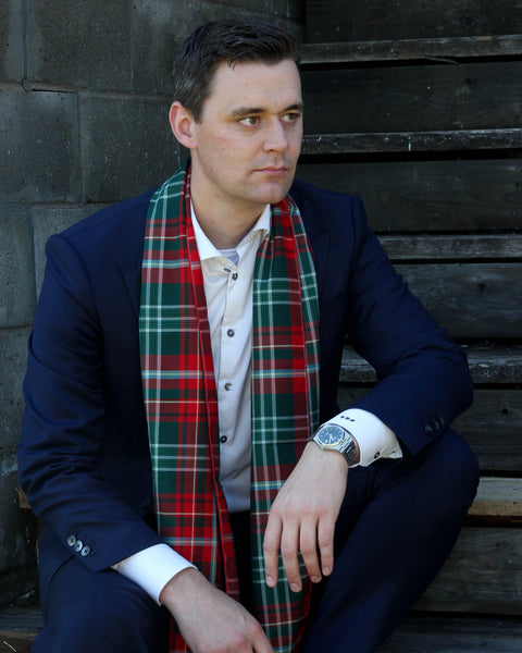 Man wearing a navy suit with a red and green plaid scarf, sitting against a wooden wall.