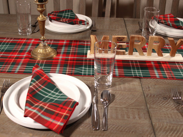 Dining table set with red and green plaid table runner, napkins, plates, glasses, and 'Merry' sign.