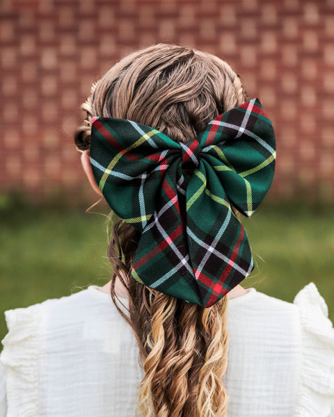 Girl with long curls wearing green and brown plaid hair bow and white blouse, in front of brick wall.