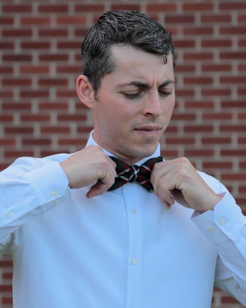 Man looking down adjusting a green plaid bow tie against a brick wall
