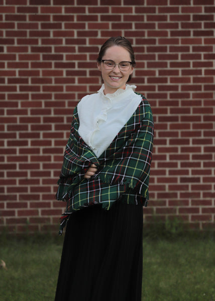 Person wearing a green plaid shawl over a white shirt and black skirt against a brick wall.
