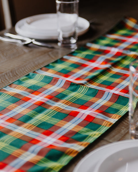 A Northwest Territories tartan plaid table runner with a festive holiday pattern in green, red, yellow, and white, laid out on a dark wooden dining table with white plates and glassware.