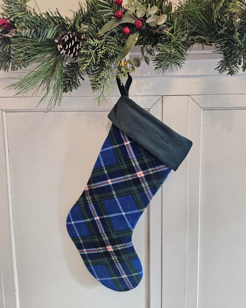 A blue and green plaid Christmas stocking with a dark green cuff hangs from a holiday garland on a white paneled door. Nova Scotia tartan fleece  stocking