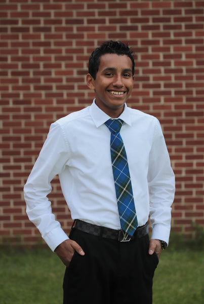 Young man wearing a nova scotia plaid neck tie with a white shirt and black dress pants standing in front of a brick wall.