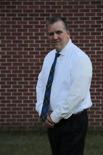 Man wearing a Nova Scotia blue and white plaid tie with a white shirt and black pants standing sideways in front of a brick wall.