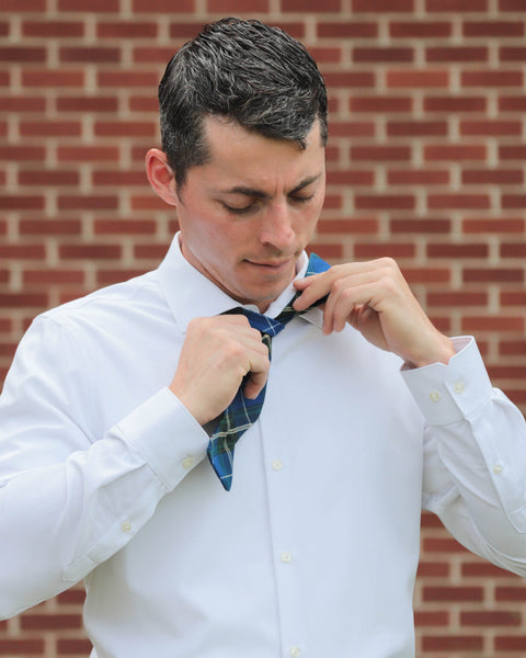 Man adjusting his Nova Scotia tartan blue plaid bow tie against a brick wall