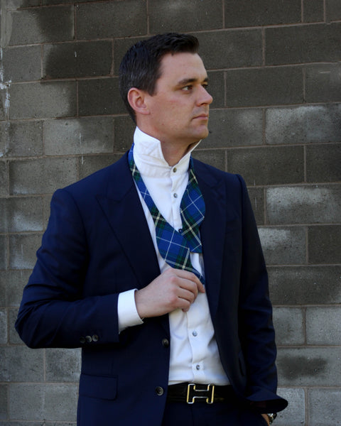 Man wearing a navy blazer, white shirt, and royal blue plaid self tie bow tie against a brick wall.