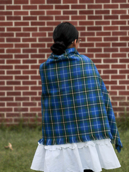 Person wearing a blue plaid shawl with a white skirt in front of a brick wall.