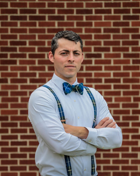 Man wearing a white shirt, blue bow tie, and plaid suspenders standing against a brick wall.