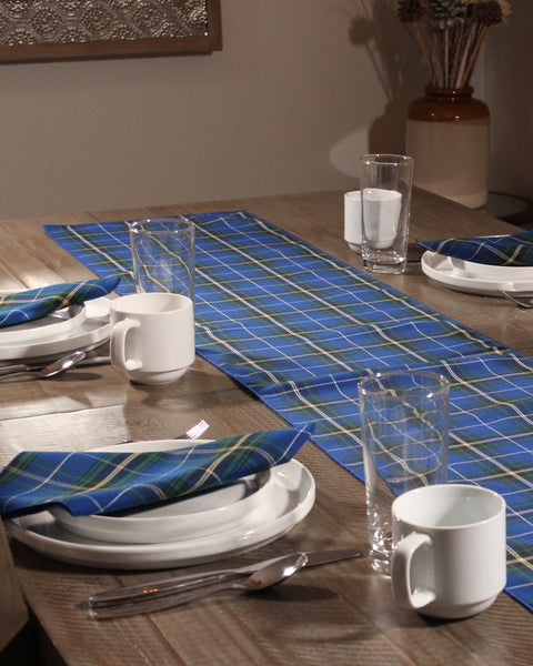 Dining table set with blue and green napkins, white plates, and glasses on a wooden table and a blue and white Nova Scotia tartan table runner.