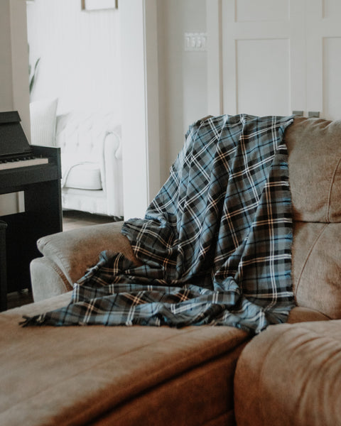 A blue and black plaid Nunavet tartan throw blanket is draped over a brown fabric sofa in a softly lit living room.