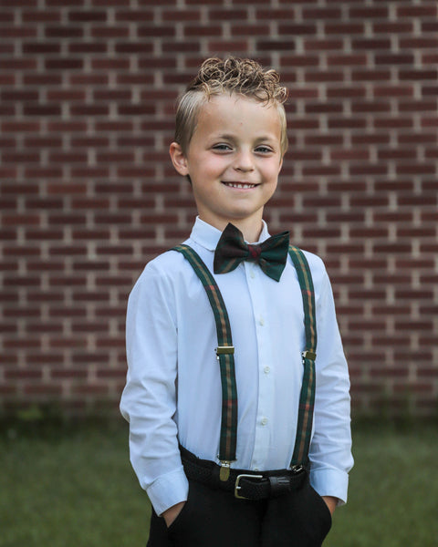 Smiling young boy wearing a bow tie and suspenders standing outdoors.