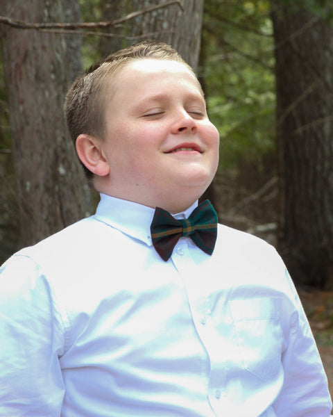 Young boy wearing a white shirt and an Ontario tartan plaid bow tie outdoors with trees in the background