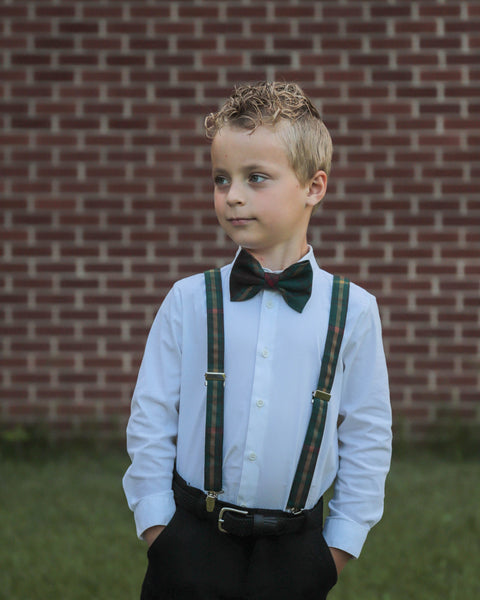 Young boy wearing a white shirt, green and gold plaid bow tie, suspenders against a brick wall.