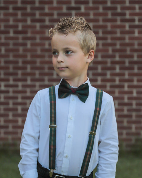 Young boy wearing a white shirt, green plaid Ontario tartan bow tie, and suspenders against a brick wall.