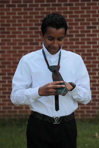Boy tying an Ontario plaid necktie wearing a white shirt and black pants.