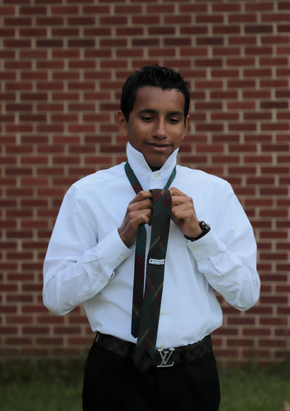 Young man tying green, gold, and red plaid necktie wearing a white shirt and black pants standing in front of a brick wall.