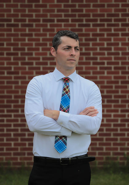 Man standing against a brick wall with arms crossed, wearing a white shirt and an Oromocto blue, red, and gold plaid tie.