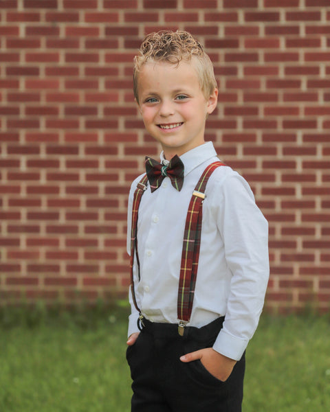 Young boy wearing a PEI tartan bow tie and suspenders standing in front of a brick wall.