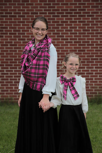 Two women wearing pink and black  plaid scarves in front of a brick wall.
