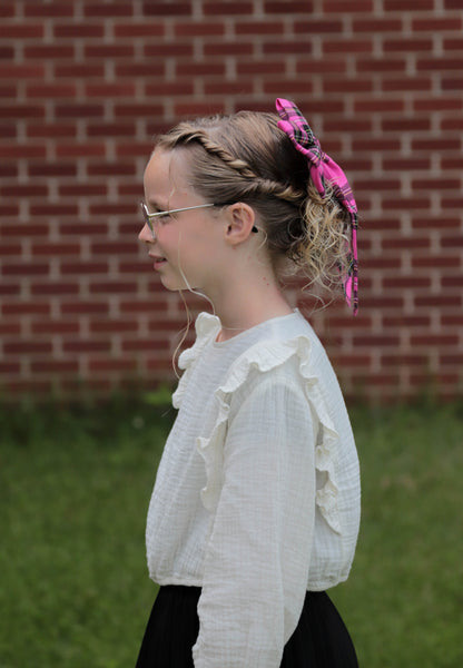 girl wearing a white blouse with ruffles and pink plaid hair bow in front of a brick wall