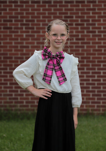 Young girl with white blouse, black skirt, pink neck bow in front of brick wall.