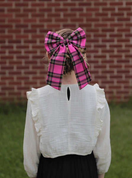 Person wearing a large pink and black plaid bow in a grassy area with a brick wall background