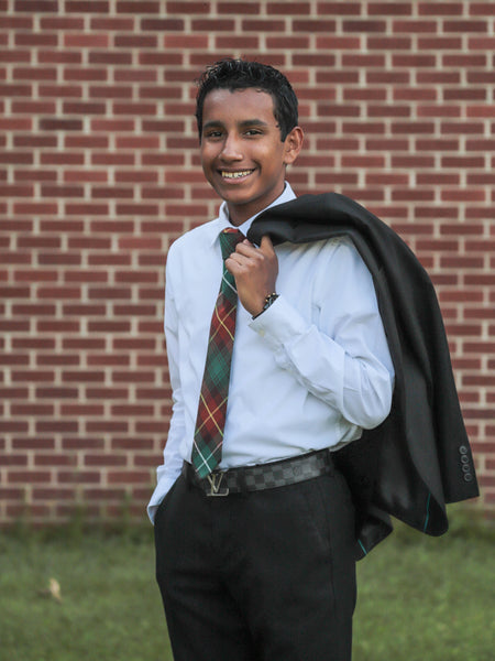 Young man in formal attire with a plaid tie and jacket over his shoulder, standing in front of a brick wall.