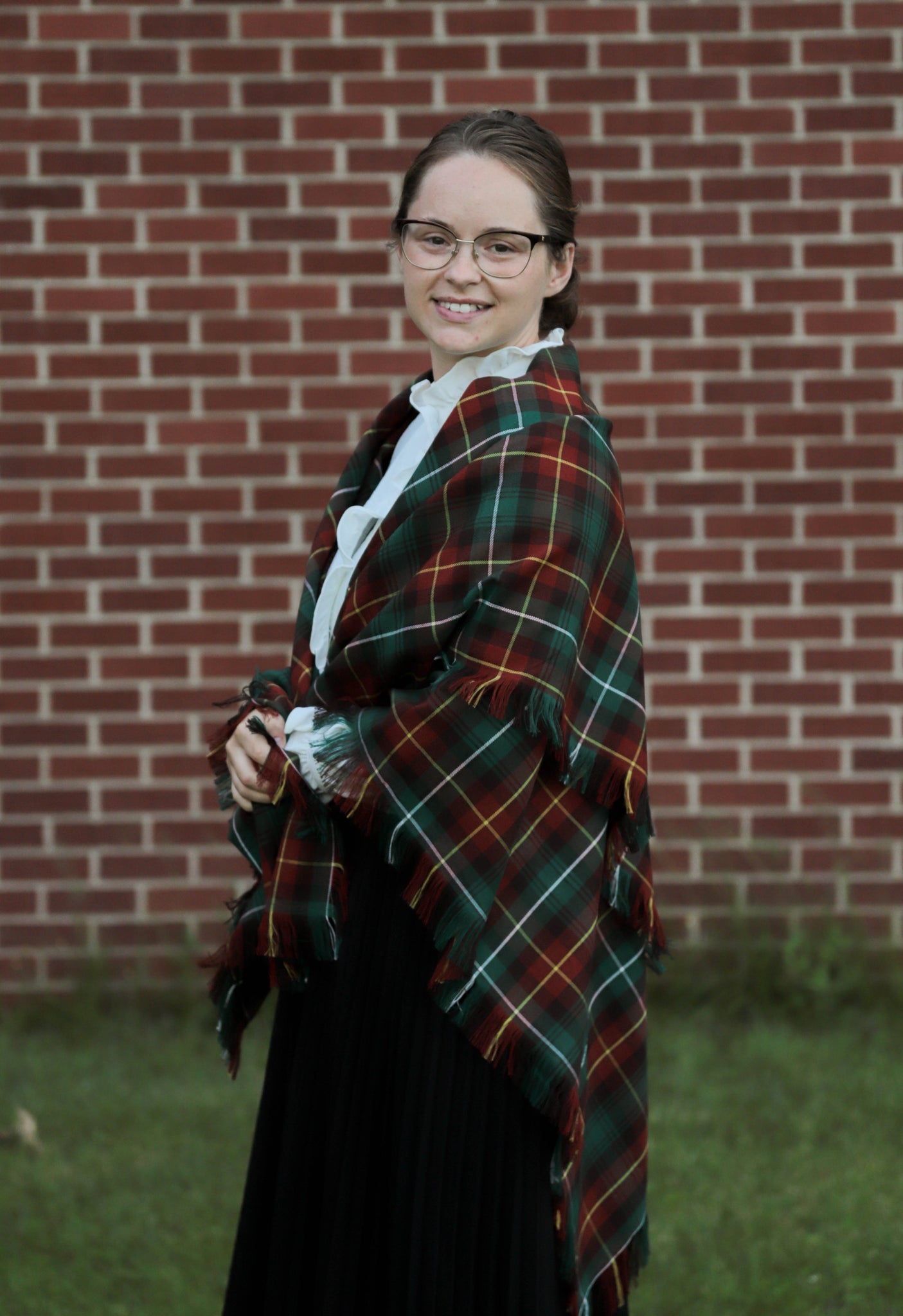 Side view of a woman wearing a green and orange plaid Prince Edward Island tartan shawl in front of a brick wall.