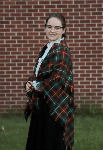 Side view of a woman wearing a green and orange plaid Prince Edward Island tartan shawl in front of a brick wall.