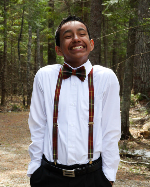 Person wearing a white shirt, red green and gold  bow tie, and suspenders standing in a forest.