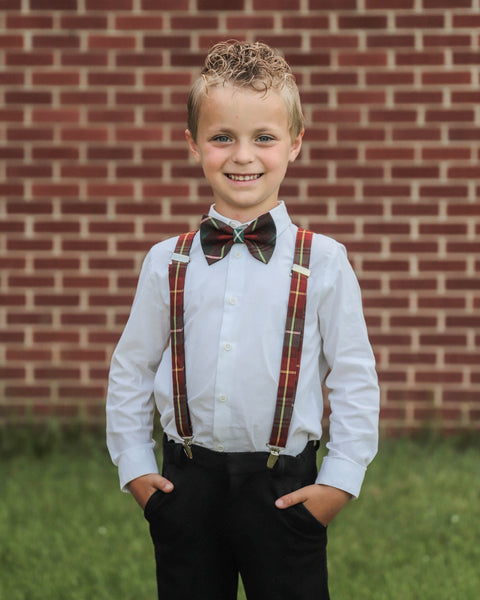 Young boy wearing a white shirt, black pants, red, green and gold PEI plaid bow tie, and suspenders standing in front of a brick wall