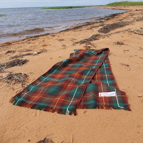 Green plaid scarf displayed on a sandy beach with ocean water and grass in the background.