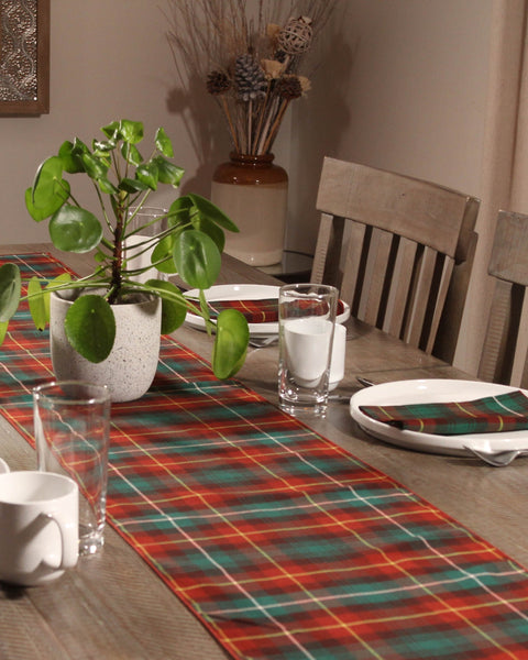 Dining table with a ggreen and rust plaid runner, glasses, plates, and a plant.