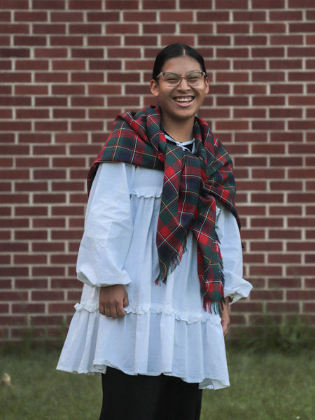 girl wearing a white top with a red and navy plaid shawl in front of a brick wall