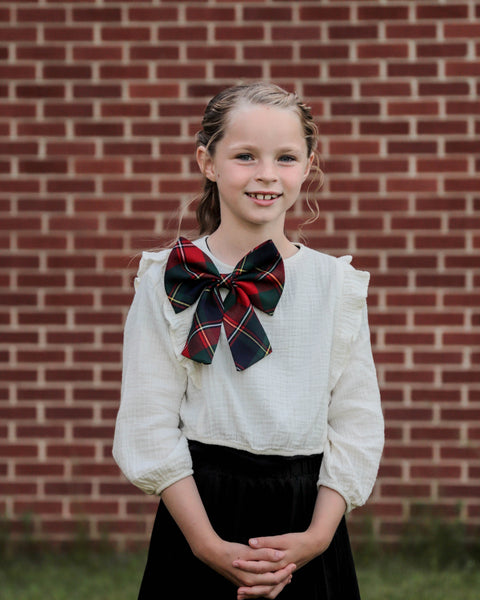 Young girl wearing a white blouse with a large tartan bow in front of a brick wall.