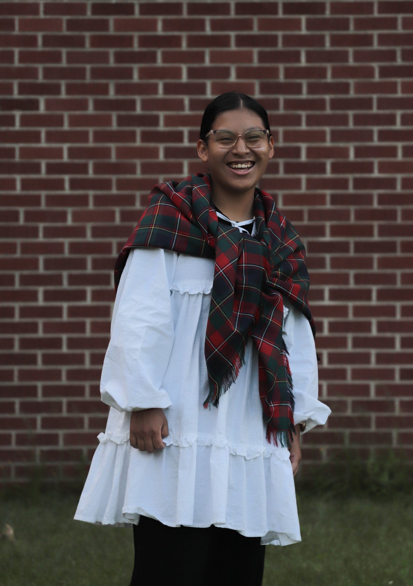 Person wearing a white dress and Quebec tartan red, green and white plaid scarf in front of a brick wall